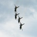 Four military jet aircraft are flying in a tight diagonal formation against an overcast sky. The jets appear to be performing an aerobatic display, flying in a stacked line-astern formation at a steep bank angle. Their afterburners appear to be engaged, visible as orange glows at the engine exhausts.