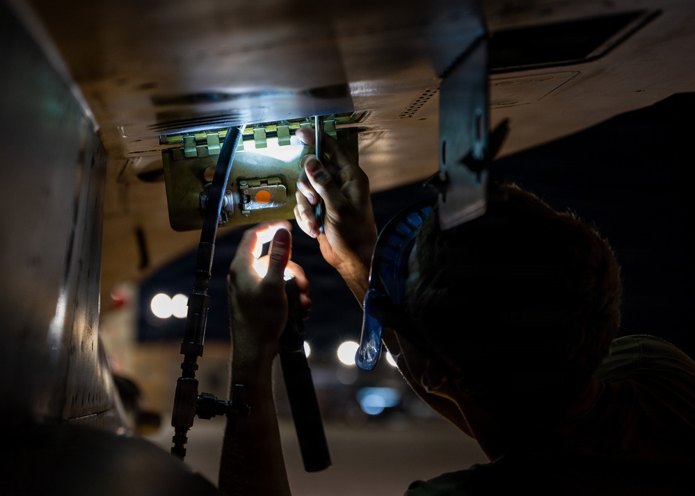 A technician or mechanic is working underneath what appears to be a military or commercial aircraft, inspecting or maintaining a mechanical component in low-light conditions. The worker is using a flashlight or penlight to illuminate the work area, which involves metal brackets, hinges, or mounting hardware on the underside of the aircraft fuselage. The image is captured in a dramatic, high-contrast style with bokeh lighting visible in the background.
