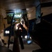 A technician or mechanic is working underneath what appears to be a military or commercial aircraft, inspecting or maintaining a mechanical component in low-light conditions. The worker is using a flashlight or penlight to illuminate the work area, which involves metal brackets, hinges, or mounting hardware on the underside of the aircraft fuselage. The image is captured in a dramatic, high-contrast style with bokeh lighting visible in the background.