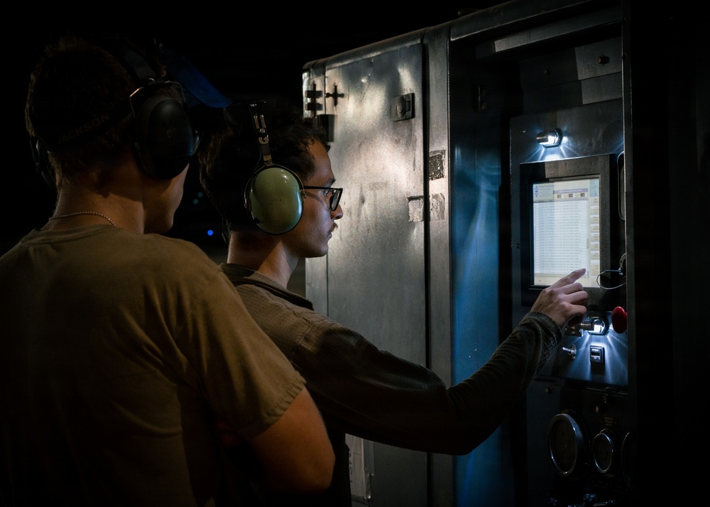 Two individuals wearing hearing protection (ear muffs) are operating or monitoring an industrial control panel in a low-light environment. One person is reaching toward the panel, which features a illuminated display screen and a red emergency stop button. The setting appears to be a military or industrial facility based on the equipment and attire.