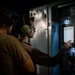 Two individuals wearing hearing protection (ear muffs) are operating or monitoring an industrial control panel in a low-light environment. One person is reaching toward the panel, which features a illuminated display screen and a red emergency stop button. The setting appears to be a military or industrial facility based on the equipment and attire.