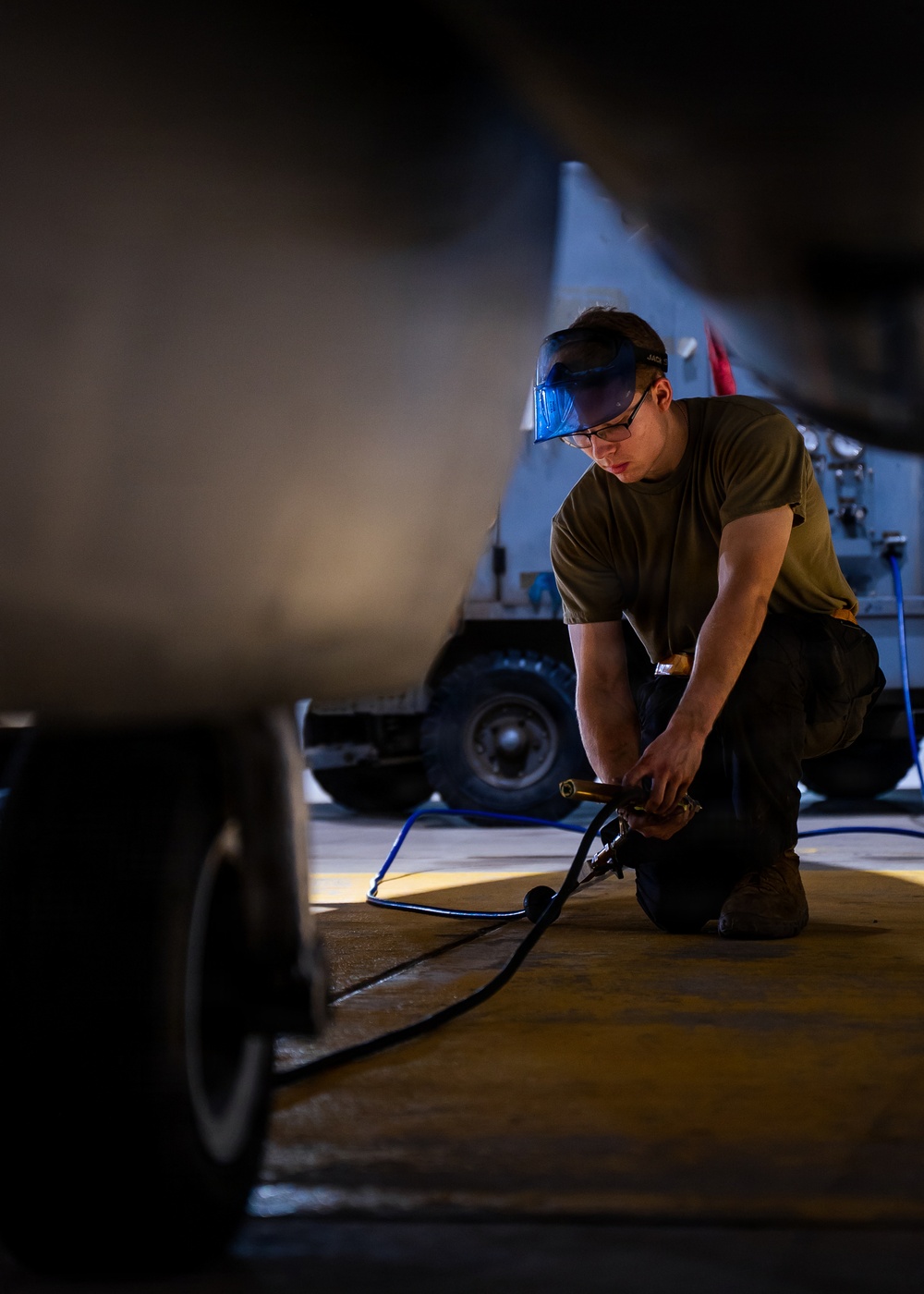 A military service member wearing an olive drab t-shirt and protective eye goggles is kneeling on a concrete floor, working with a pneumatic or hydraulic tool connected to a blue hose. The image is captured from a low angle between two large vehicles, likely aircraft or military equipment, in what appears to be a hangar or flight line environment. A military support vehicle is visible in the background.