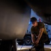 A military service member wearing an olive drab t-shirt and protective eye goggles is kneeling on a concrete floor, working with a pneumatic or hydraulic tool connected to a blue hose. The image is captured from a low angle between two large vehicles, likely aircraft or military equipment, in what appears to be a hangar or flight line environment. A military support vehicle is visible in the background.