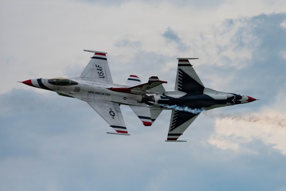 Two United States Air Force Thunderbirds F-16 Fighting Falcon jets are performing a close-formation aerobatic maneuver, appearing to fly in opposite orientations — one inverted relative to the other. The aircraft display the iconic Thunderbirds red, white, and blue livery with 'USAF' and 'United States Air Force' markings clearly visible. One jet is emitting a white smoke trail, characteristic of Thunderbirds airshow performances.