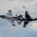 Two United States Air Force Thunderbirds F-16 Fighting Falcon jets are performing a close-formation aerobatic maneuver, appearing to fly in opposite orientations — one inverted relative to the other. The aircraft display the iconic Thunderbirds red, white, and blue livery with 'USAF' and 'United States Air Force' markings clearly visible. One jet is emitting a white smoke trail, characteristic of Thunderbirds airshow performances.