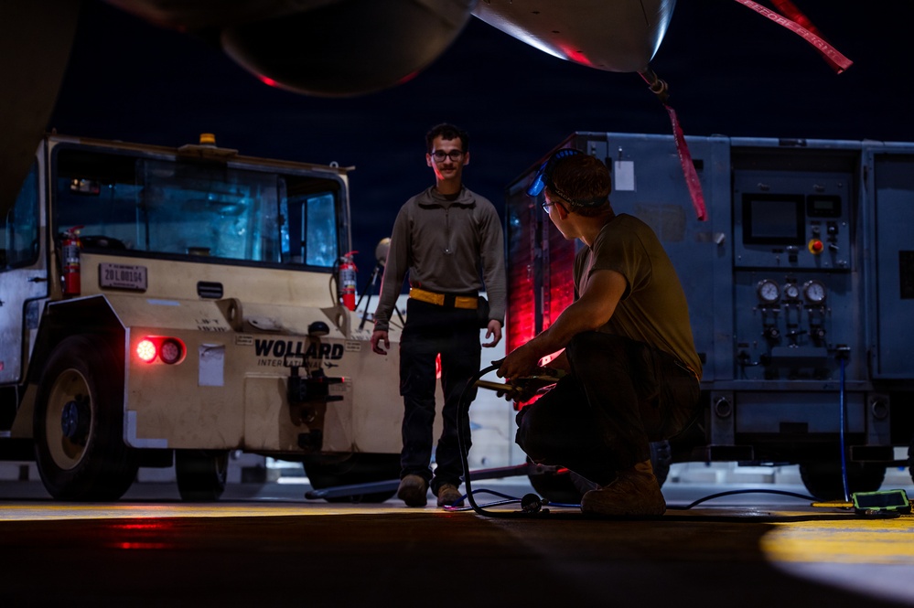 A nighttime aircraft maintenance or ground operations scene on a flight line or tarmac. Two personnel are working near the underside of an aircraft, with one crouching and handling equipment while the other stands observing. Ground support equipment, including a Wollard International tug vehicle and what appears to be a power or hydraulic cart, are visible in the background.