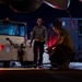 A nighttime aircraft maintenance or ground operations scene on a flight line or tarmac. Two personnel are working near the underside of an aircraft, with one crouching and handling equipment while the other stands observing. Ground support equipment, including a Wollard International tug vehicle and what appears to be a power or hydraulic cart, are visible in the background.