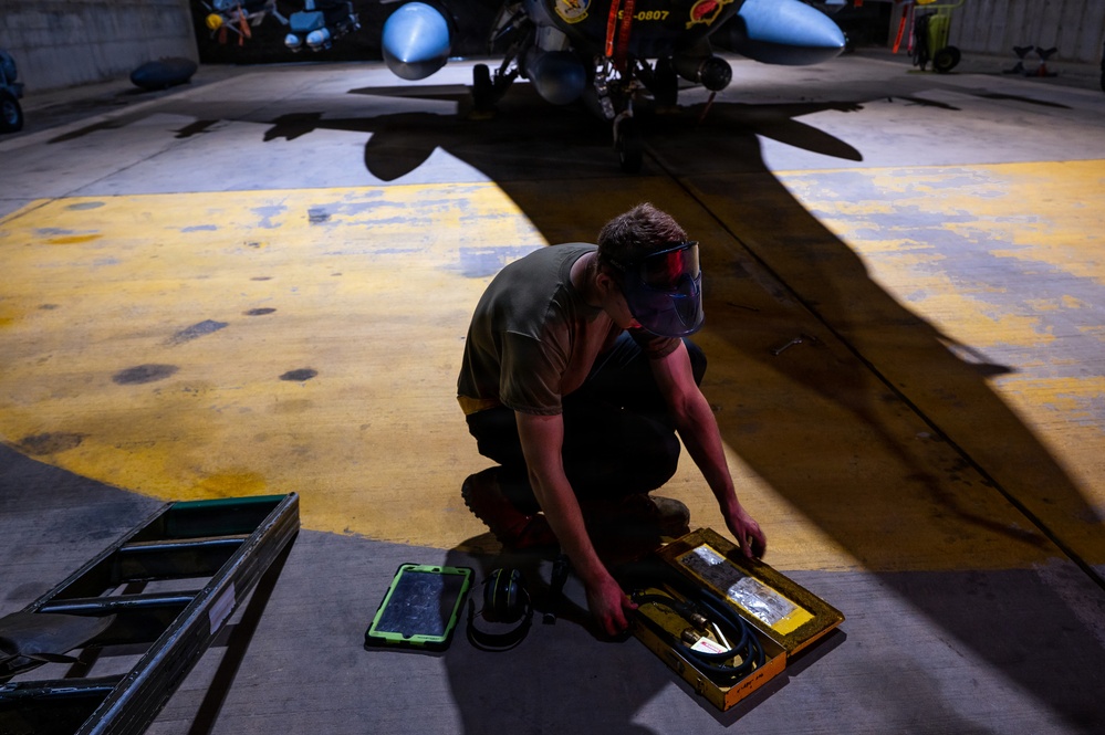 A technician or maintenance worker is crouching on a hangar floor, working with a yellow tool or equipment case. The setting appears to be a military aircraft hangar, with a jet aircraft visible in the background bearing the tail number 0807. The worker is wearing a protective face shield and olive/military-colored clothing.