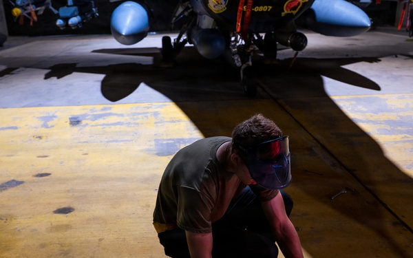 A technician or maintenance worker is crouching on a hangar floor, working with a yellow tool or equipment case. The setting appears to be a military aircraft hangar, with a jet aircraft visible in the background bearing the tail number 0807. The worker is wearing a protective face shield and olive/military-colored clothing.