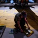 A technician or maintenance worker is crouching on a hangar floor, working with a yellow tool or equipment case. The setting appears to be a military aircraft hangar, with a jet aircraft visible in the background bearing the tail number 0807. The worker is wearing a protective face shield and olive/military-colored clothing.