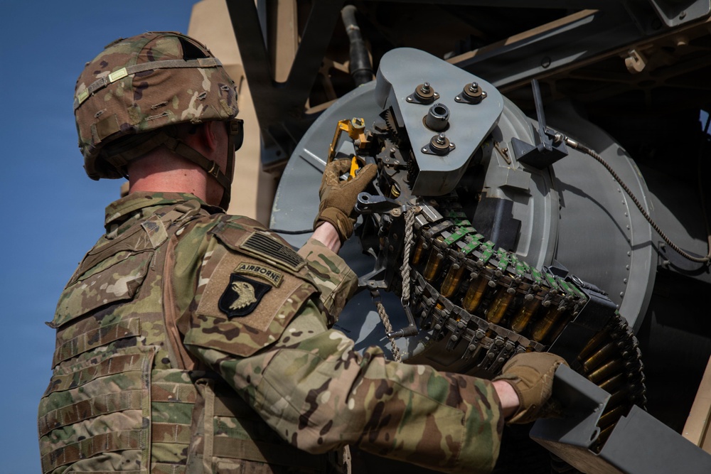 A U.S. Army soldier from the 101st Airborne Division is loading or maintaining an ammunition belt into a vehicle-mounted weapon system. The soldier is wearing MultiCam (OCP) camouflage uniform with an 'Airborne' tab and the 101st Airborne Division patch visible on the left shoulder. The image is taken from behind and slightly above, showing the soldier working on what appears to be a crew-served weapon mounted on a military vehicle.