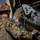 A U.S. Army soldier from the 101st Airborne Division is loading or maintaining an ammunition belt into a vehicle-mounted weapon system. The soldier is wearing MultiCam (OCP) camouflage uniform with an 'Airborne' tab and the 101st Airborne Division patch visible on the left shoulder. The image is taken from behind and slightly above, showing the soldier working on what appears to be a crew-served weapon mounted on a military vehicle.