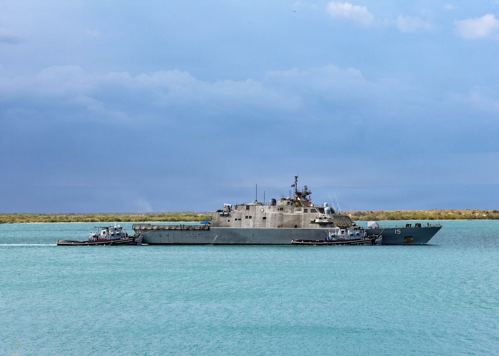 A U.S. Navy Littoral Combat Ship bearing hull number 15 is underway in turquoise coastal waters, assisted by two tugboats on either side. The vessel displays the characteristic low-profile, angular design of the Freedom-class or Independence-class LCS. The scene appears to be a port departure or arrival maneuver in a tropical or semi-arid coastal environment.