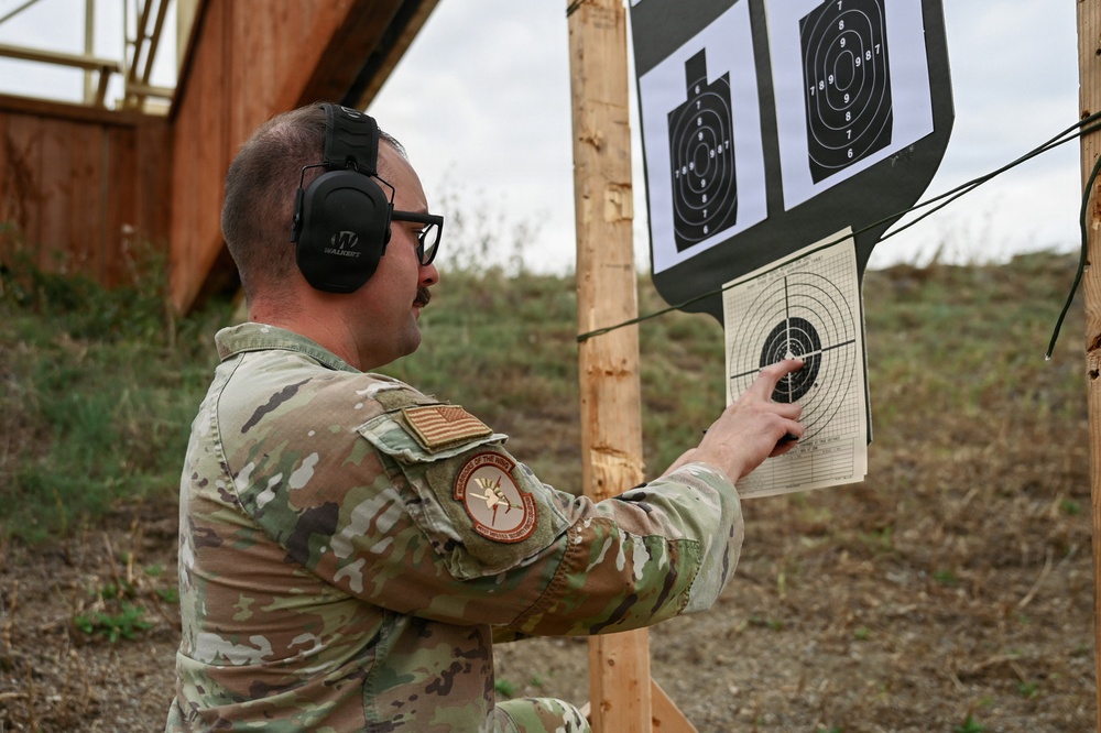 A military service member in MultiCam/OCP camouflage uniform is examining shooting targets at an outdoor firing range. The individual is pointing at a circular bullseye target, likely assessing shot placement and accuracy. The setting appears to be a standard military qualification range with multiple target types mounted on wooden posts.