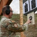 A military service member in MultiCam/OCP camouflage uniform is examining shooting targets at an outdoor firing range. The individual is pointing at a circular bullseye target, likely assessing shot placement and accuracy. The setting appears to be a standard military qualification range with multiple target types mounted on wooden posts.