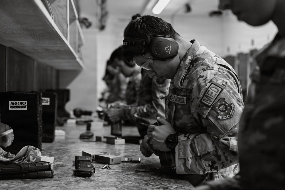 A U.S. Air Force service member wearing OCP (Operational Camouflage Pattern) uniform is at an indoor shooting range, focused on handling or loading a firearm at a shooting bench. The individual is equipped with hearing protection (earmuffs) and eye protection (safety glasses), indicating active range safety protocols. Several other uniformed personnel are visible in the background performing similar activities along the range line.