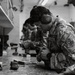 A U.S. Air Force service member wearing OCP (Operational Camouflage Pattern) uniform is at an indoor shooting range, focused on handling or loading a firearm at a shooting bench. The individual is equipped with hearing protection (earmuffs) and eye protection (safety glasses), indicating active range safety protocols. Several other uniformed personnel are visible in the background performing similar activities along the range line.