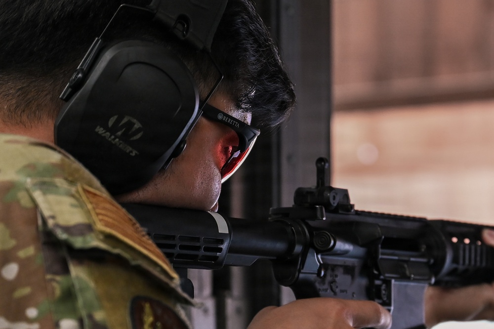 A military or law enforcement personnel is aiming a rifle at a shooting range. The individual is wearing proper personal protective equipment including hearing protection and eye protection. The image is a close-up shot focusing on the shooter's head and the rifle.