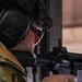 A military or law enforcement personnel is aiming a rifle at a shooting range. The individual is wearing proper personal protective equipment including hearing protection and eye protection. The image is a close-up shot focusing on the shooter's head and the rifle.