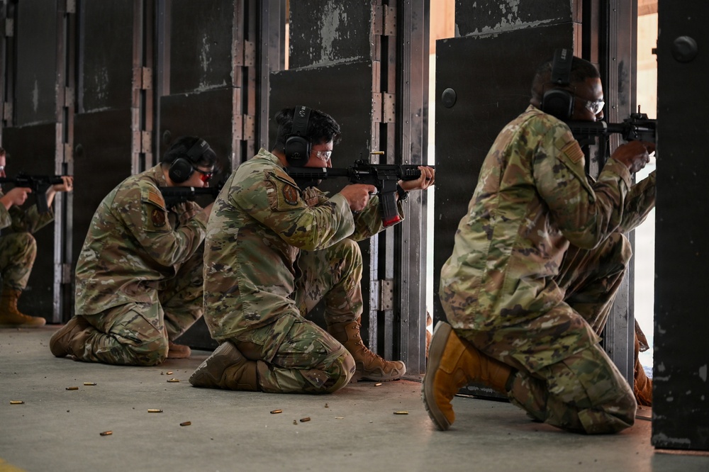 Multiple military personnel in OCP (Operational Camouflage Pattern) uniforms are conducting weapons qualification or training at an indoor shooting range. The soldiers are in kneeling positions, aiming rifles downrange through designated shooting lanes separated by metal dividers. Spent brass casings are visible on the concrete floor, indicating active live-fire exercise.