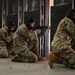 Multiple military personnel in OCP (Operational Camouflage Pattern) uniforms are conducting weapons qualification or training at an indoor shooting range. The soldiers are in kneeling positions, aiming rifles downrange through designated shooting lanes separated by metal dividers. Spent brass casings are visible on the concrete floor, indicating active live-fire exercise.