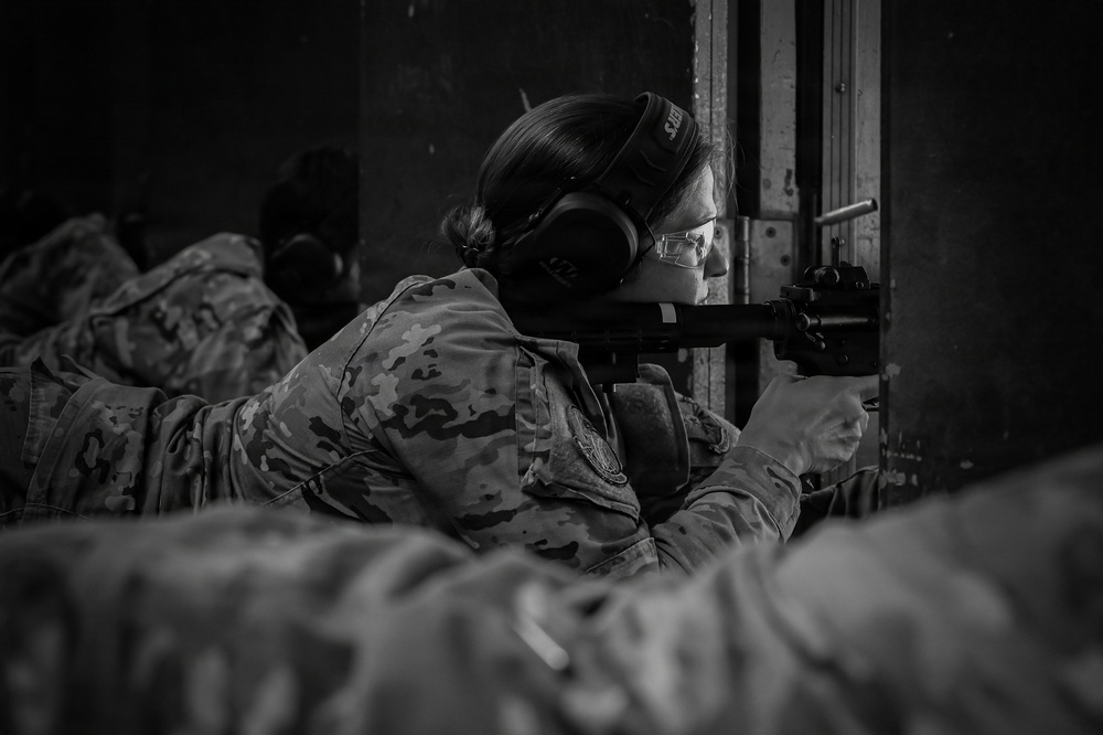 A black-and-white photograph showing a female military or law enforcement personnel in camouflage uniform aiming a rifle at a shooting range. She is wearing hearing protection earmuffs and safety glasses, indicating a formal training or qualification exercise. Additional personnel in camouflage uniforms are visible in the background and foreground.