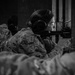 A black-and-white photograph showing a female military or law enforcement personnel in camouflage uniform aiming a rifle at a shooting range. She is wearing hearing protection earmuffs and safety glasses, indicating a formal training or qualification exercise. Additional personnel in camouflage uniforms are visible in the background and foreground.