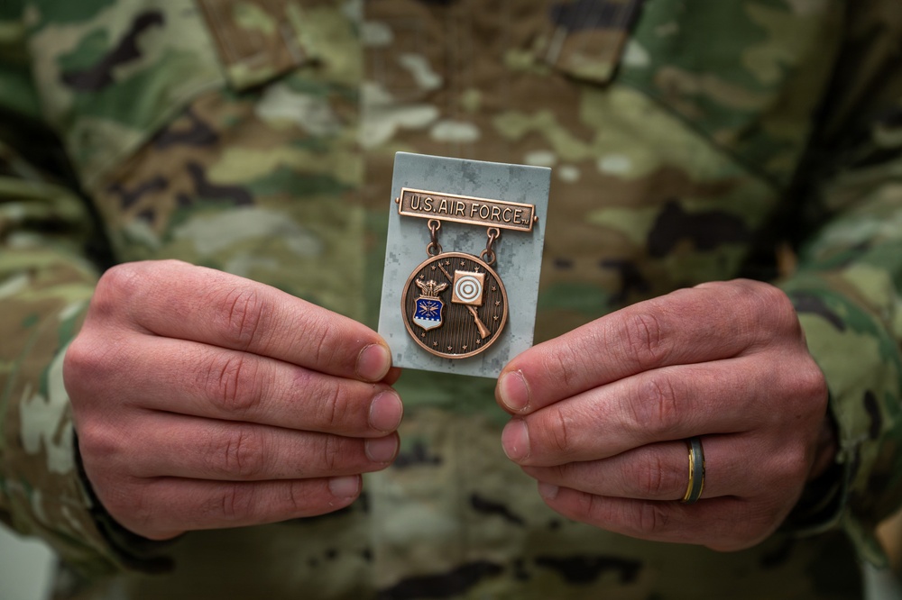 A person wearing OCP (Operational Camouflage Pattern) military uniform is holding a U.S. Air Force marksmanship badge. The bronze medal features the Air Force shield emblem on the left and a target/rifle design on the right, suspended from a bar engraved 'U.S. AIR FORCE.' The individual is wearing a wedding ring on their left hand.