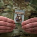 A person wearing OCP (Operational Camouflage Pattern) military uniform is holding a U.S. Air Force marksmanship badge. The bronze medal features the Air Force shield emblem on the left and a target/rifle design on the right, suspended from a bar engraved 'U.S. AIR FORCE.' The individual is wearing a wedding ring on their left hand.