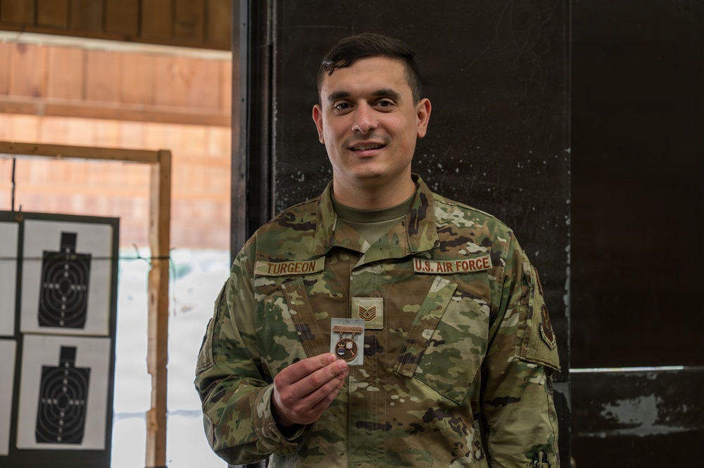 A U.S. Air Force Technical Sergeant named Turgeon is posing at what appears to be an indoor shooting range, holding a small medal or badge. He is wearing OCP (Operational Camouflage Pattern) uniform with visible name tape and branch tape. The setting includes paper shooting targets visible in the background.