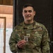 A U.S. Air Force Technical Sergeant named Turgeon is posing at what appears to be an indoor shooting range, holding a small medal or badge. He is wearing OCP (Operational Camouflage Pattern) uniform with visible name tape and branch tape. The setting includes paper shooting targets visible in the background.
