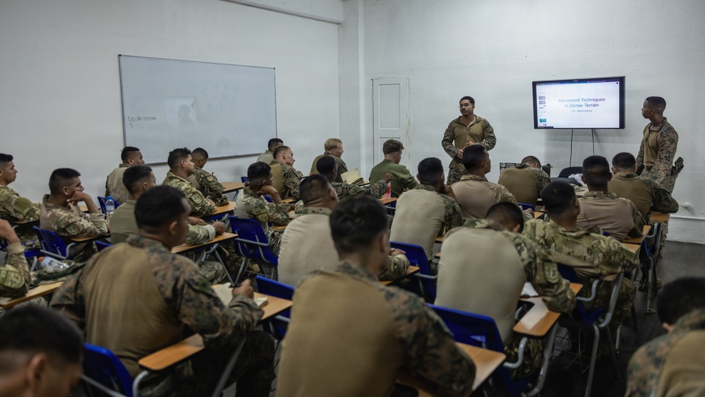 Military personnel are attending a classroom instruction session on 'Movement Techniques in Dense Terrain,' as displayed on a wall-mounted screen. Two instructors stand at the front of the room while approximately 20-30 soldiers/marines in camouflage uniforms sit at individual desks taking notes. The setting appears to be a formal military training environment with a whiteboard and presentation display.