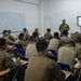 Military personnel are attending a classroom instruction session on 'Movement Techniques in Dense Terrain,' as displayed on a wall-mounted screen. Two instructors stand at the front of the room while approximately 20-30 soldiers/marines in camouflage uniforms sit at individual desks taking notes. The setting appears to be a formal military training environment with a whiteboard and presentation display.