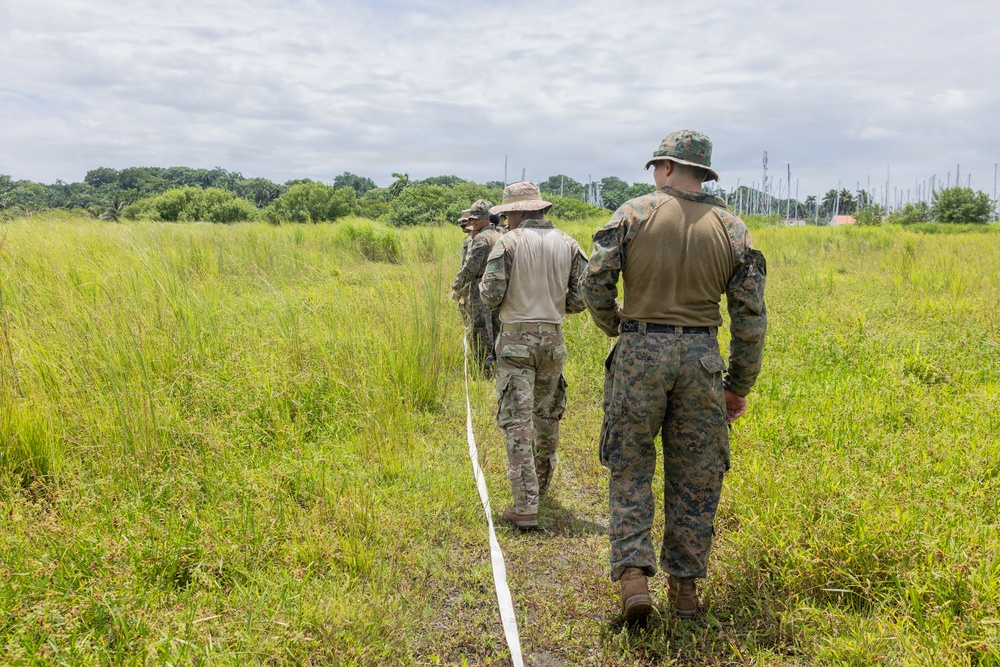 Three military personnel in camouflage uniforms are walking through a grassy field, following a white rope or line on the ground. They appear to be conducting a field exercise or survey operation in a tropical or subtropical environment. The personnel are wearing different camouflage patterns, suggesting possible joint or multinational military activity.