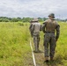 Three military personnel in camouflage uniforms are walking through a grassy field, following a white rope or line on the ground. They appear to be conducting a field exercise or survey operation in a tropical or subtropical environment. The personnel are wearing different camouflage patterns, suggesting possible joint or multinational military activity.