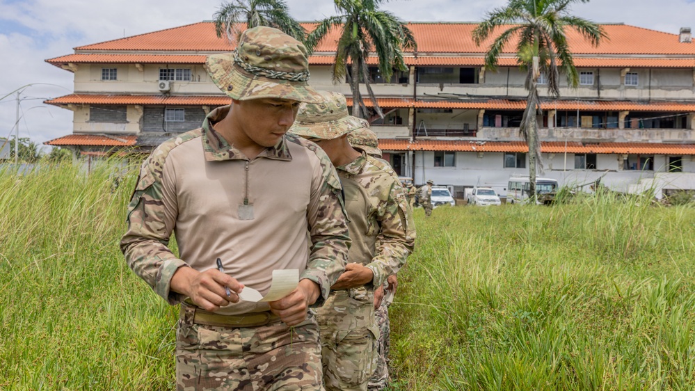 Two military personnel in MultiCam/OCP camouflage uniforms and boonie hats are reviewing a small document or notepad in an overgrown field. They are positioned in front of a large multi-story building with a terracotta roof and palm trees, suggesting a tropical location. Additional personnel and vehicles are visible in the background.