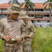 Two military personnel in MultiCam/OCP camouflage uniforms and boonie hats are reviewing a small document or notepad in an overgrown field. They are positioned in front of a large multi-story building with a terracotta roof and palm trees, suggesting a tropical location. Additional personnel and vehicles are visible in the background.