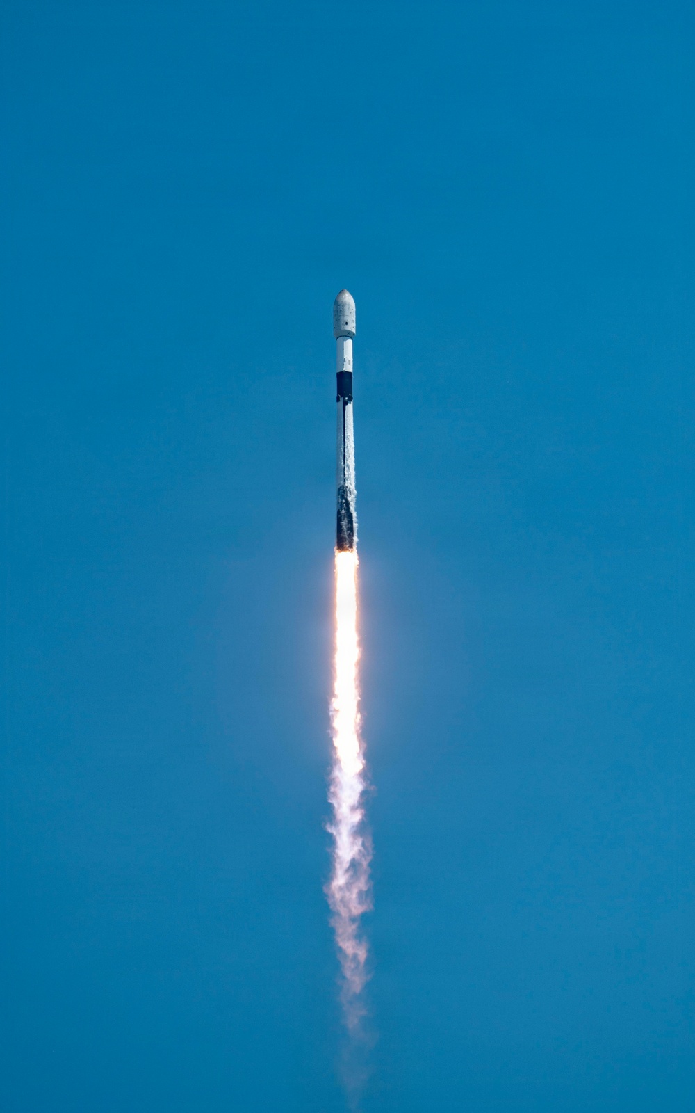A rocket is captured mid-flight ascending vertically against a clear blue sky. The rocket displays a white and black color scheme with a visible payload fairing at the top, consistent with a Falcon 9-style vehicle. A bright orange flame and white exhaust plume trail beneath the vehicle, indicating active first-stage engine burn.