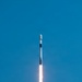 A rocket is captured mid-flight ascending vertically against a clear blue sky. The rocket displays a white and black color scheme with a visible payload fairing at the top, consistent with a Falcon 9-style vehicle. A bright orange flame and white exhaust plume trail beneath the vehicle, indicating active first-stage engine burn.