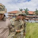Three military personnel in camouflage uniforms and boonie hats are walking through tall grass in a line formation, appearing to conduct a search or survey operation. They are holding writing instruments and papers, suggesting documentation or data collection activity. The setting appears to be a tropical location with palm trees and a multi-story building with an orange roof visible in the background.