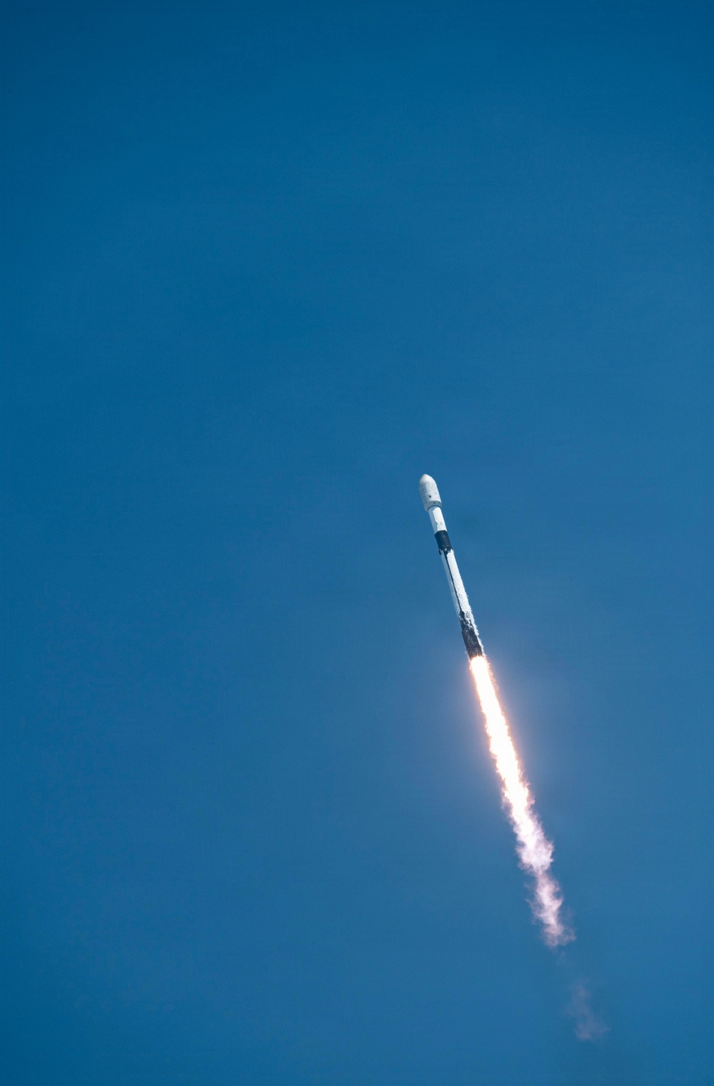 A rocket is captured mid-flight ascending through a clear blue sky at a diagonal angle. A bright plume of fire and white exhaust trail extends behind the vehicle, indicating active engine burn. The rocket appears to be a two-stage vehicle with a white payload fairing at the top.