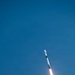 A rocket is captured mid-flight ascending through a clear blue sky at a diagonal angle. A bright plume of fire and white exhaust trail extends behind the vehicle, indicating active engine burn. The rocket appears to be a two-stage vehicle with a white payload fairing at the top.