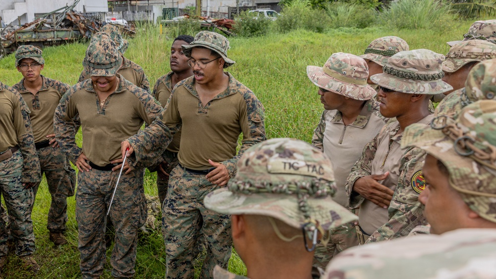 A group of military personnel in two distinct uniform types are gathered outdoors in a grassy area near what appears to be a debris field or scrapyard. One individual wearing MARPAT (Marine digital camouflage) appears to be conducting a briefing or instruction session, holding a pointer or stick. The group includes personnel in both MARPAT uniforms and MultiCam/OCP-style uniforms, suggesting a joint or multinational training exercise.
