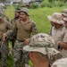 A group of military personnel in two distinct uniform types are gathered outdoors in a grassy area near what appears to be a debris field or scrapyard. One individual wearing MARPAT (Marine digital camouflage) appears to be conducting a briefing or instruction session, holding a pointer or stick. The group includes personnel in both MARPAT uniforms and MultiCam/OCP-style uniforms, suggesting a joint or multinational training exercise.