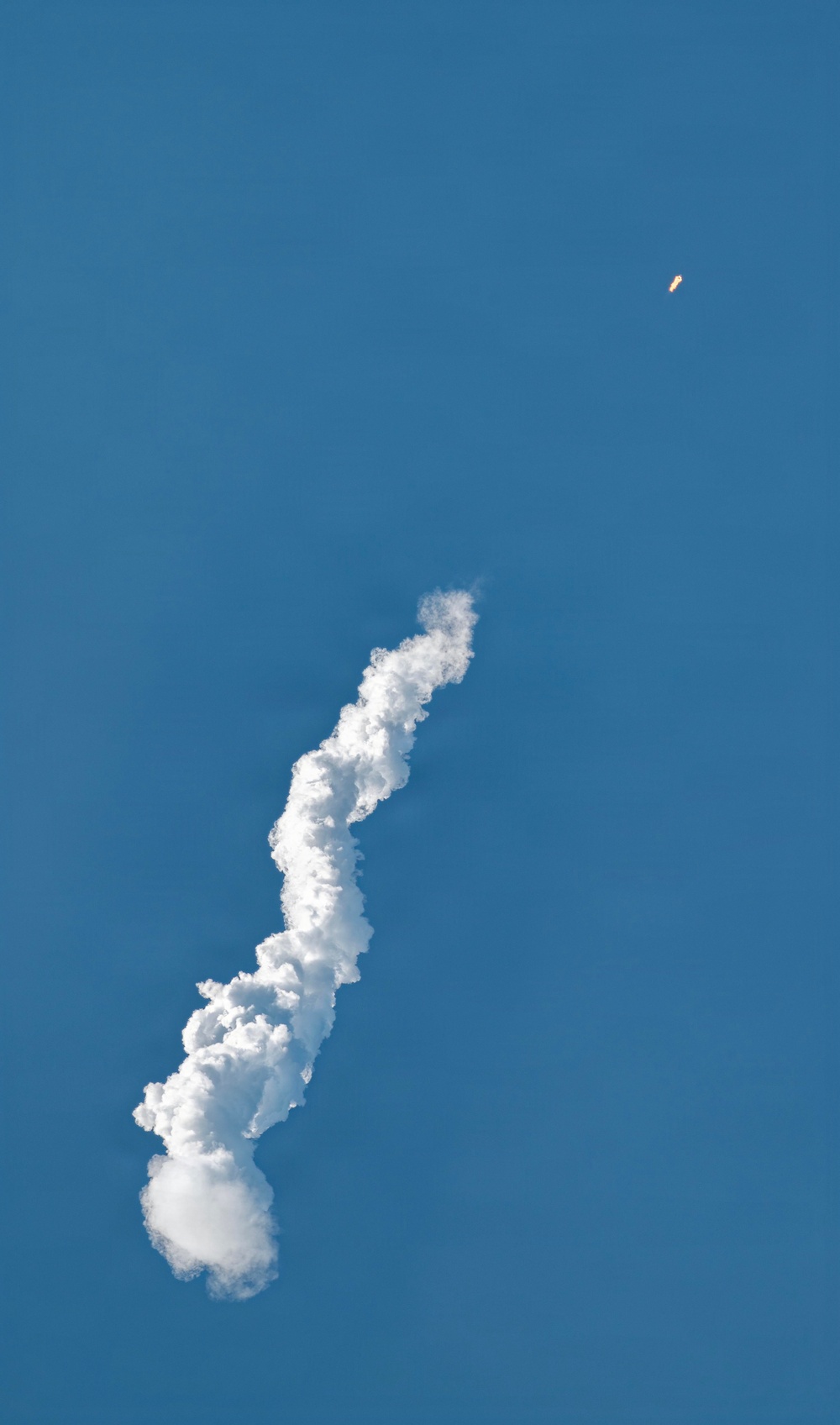The image shows a rocket ascending through a clear blue sky, leaving a thick, billowing white exhaust/smoke trail behind it. The rocket itself is visible in the upper right portion of the frame as a small orange-yellow object, having traveled a significant distance from its launch point. The smoke trail curves and widens as it descends toward the lower left of the frame, indicating the rocket's flight path.