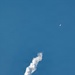The image shows a rocket ascending through a clear blue sky, leaving a thick, billowing white exhaust/smoke trail behind it. The rocket itself is visible in the upper right portion of the frame as a small orange-yellow object, having traveled a significant distance from its launch point. The smoke trail curves and widens as it descends toward the lower left of the frame, indicating the rocket's flight path.