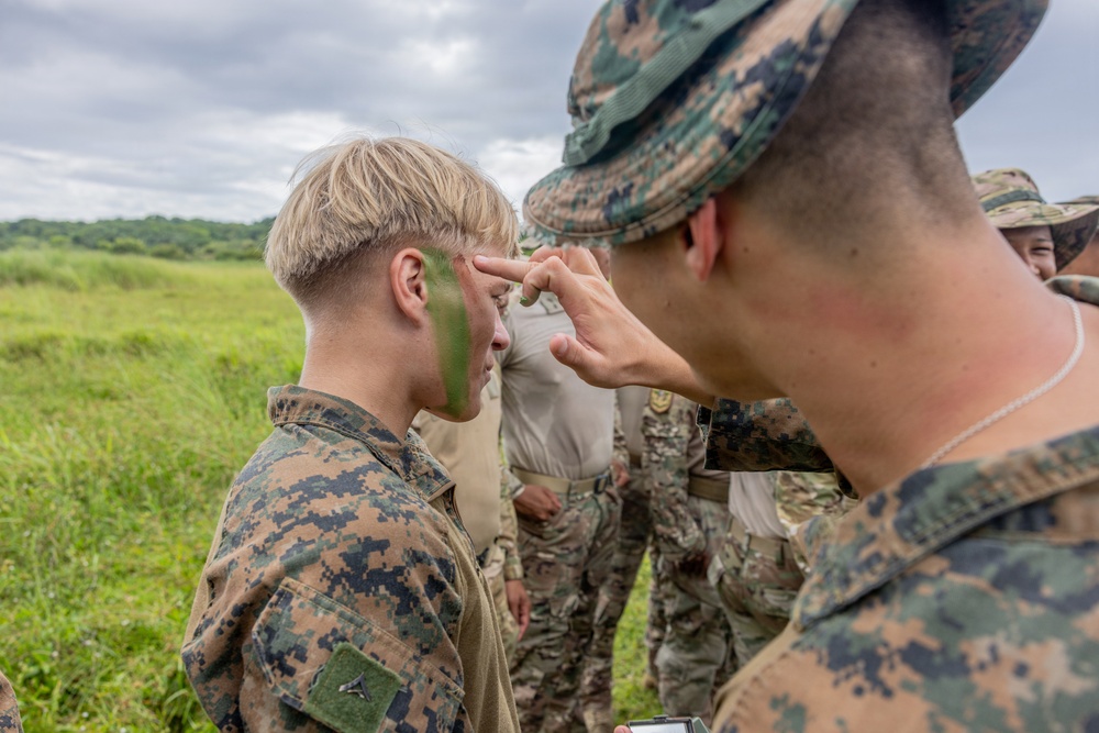 Military personnel in camouflage uniforms are applying green face paint (camouflage paint) to each other in an outdoor field setting. The scene appears to be part of a field training exercise or pre-mission preparation. Multiple service members wearing MARPAT (Marine pattern) and other camouflage uniforms are visible in the background.