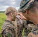 Military personnel in camouflage uniforms are applying green face paint (camouflage paint) to each other in an outdoor field setting. The scene appears to be part of a field training exercise or pre-mission preparation. Multiple service members wearing MARPAT (Marine pattern) and other camouflage uniforms are visible in the background.