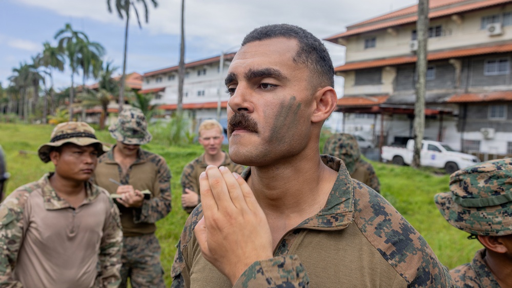 A group of military personnel in camouflage uniforms are gathered outdoors near multi-story buildings surrounded by tropical vegetation. The central figure, wearing MARPAT (Marine digital camouflage) and camouflage face paint, appears to be in the process of applying or adjusting face paint. The setting suggests a tropical or subtropical military installation.