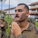 A group of military personnel in camouflage uniforms are gathered outdoors near multi-story buildings surrounded by tropical vegetation. The central figure, wearing MARPAT (Marine digital camouflage) and camouflage face paint, appears to be in the process of applying or adjusting face paint. The setting suggests a tropical or subtropical military installation.